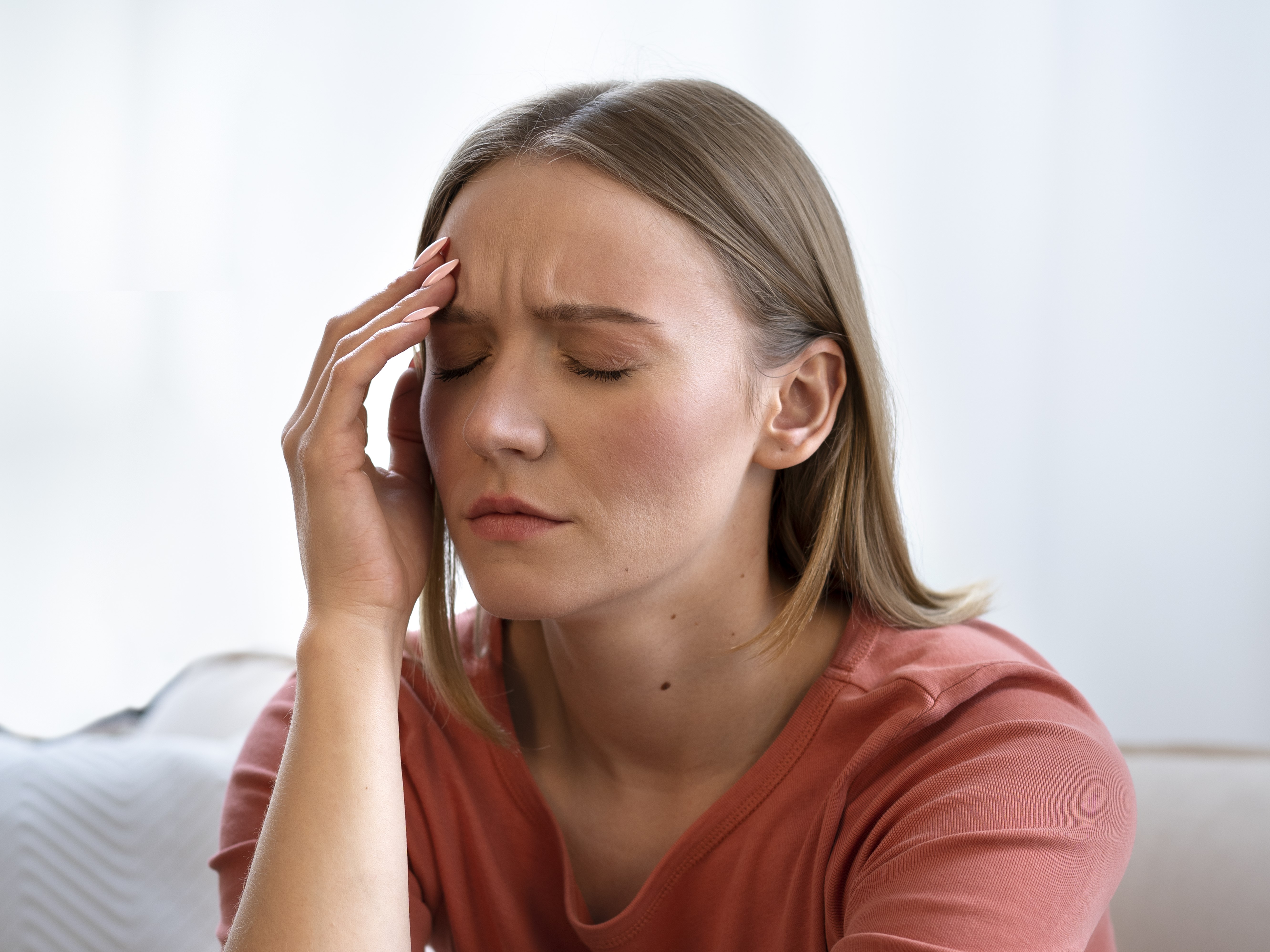A imagem mostra uma mulher com expressão de desconforto, olhos fechados e a mão na cabeça, sugerindo dor de cabeça ou cansaço, o que pode estar relacionado a sintomas de desequilíbrio hormonal, como a baixa de progesterona.
