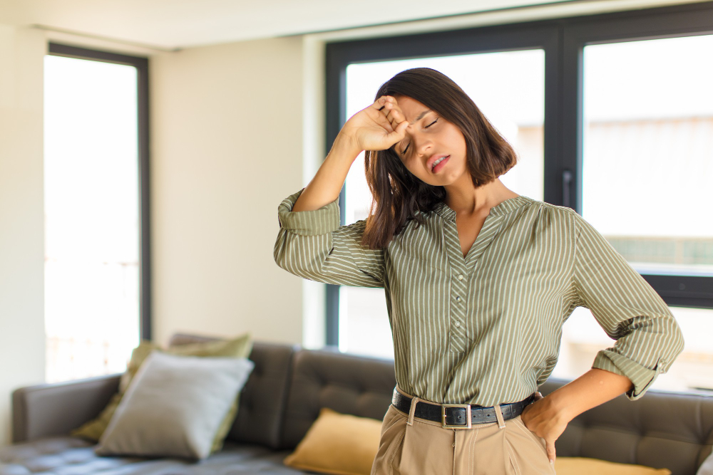 A imagem mostra uma mulher com expressão de cansaço e desconforto, vestindo uma camisa listrada verde e bege e calça bege. Ela está em um ambiente iluminado, aparentemente uma sala de estar, com um sofá escuro e almofadas ao fundo. A mulher tem a mão esquerda apoiada na cintura e a direita na testa, demonstrando exaustão. Sua pele parece pálida, e sua postura indica fadiga, sintomas comuns de anemia.