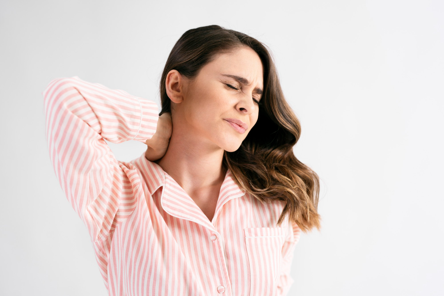 A imagem mostra uma mulher com a mão no pescoço que aparenta estar com dor. Tem cabelos pouco abaixo do ombro, castanhos e veste uma camisa rosa claro com listras brancas.