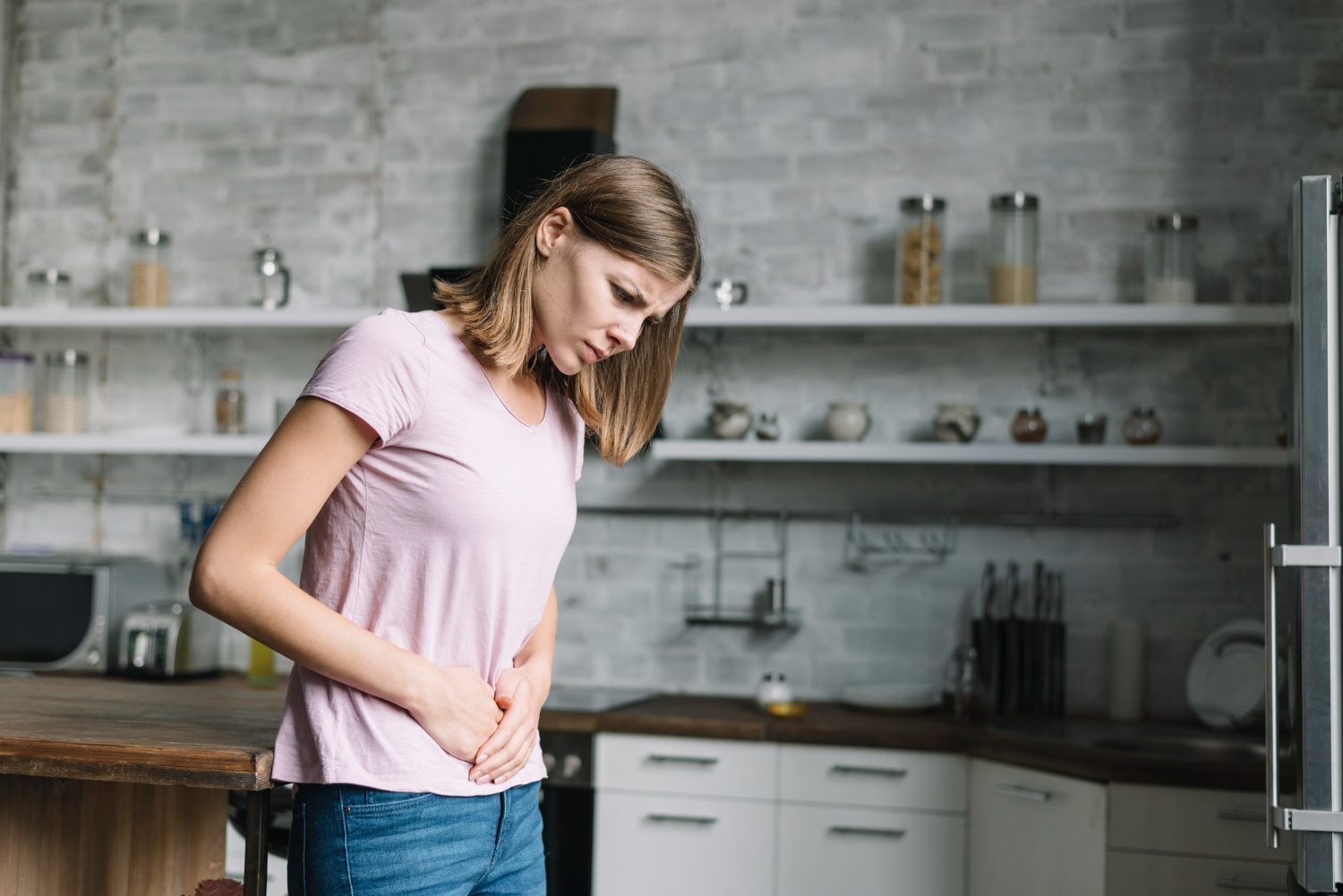 A imagem mostra uma mulher, de cabelos loiros na altura do ombro,  que está em uma cozinha,  ela está com as duas mãos na barriga e tem uma expressão de dor. Ela veste camiseta rosa claro e calça jeans.