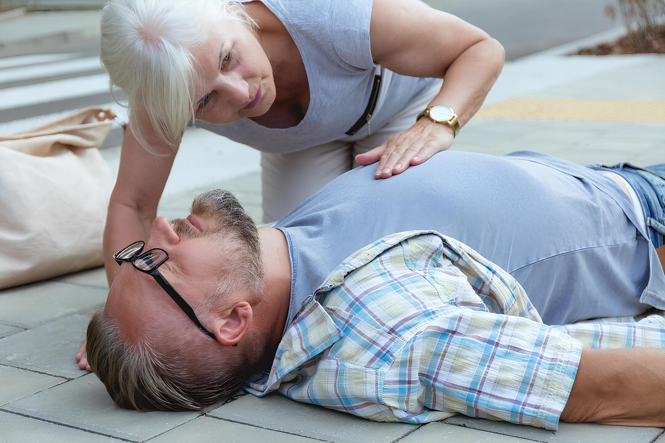 A imagem mostra um homem desmaiado no chão, ele está de óculos, camiseta azul e camisa xadrez azul. Uma mulher de cabelos brancos verifica se ele está respirando.