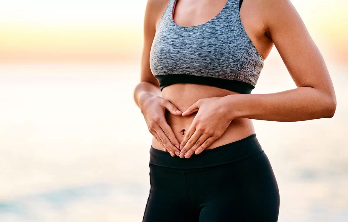 Mulher em roupas esportivas fazendo um gesto de coração com as mãos sobre o abdômen, simbolizando cuidado e atenção à saúde intestinal, ao ar livre com um cenário de pôr do sol ao fundo..