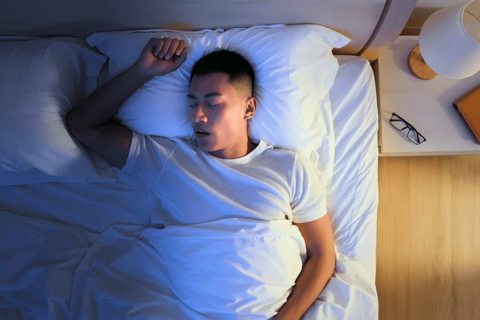 Homem dormindo deitado em uma cama durante a noite, com a boca aberta e sinais que remetem à apneia obstrutiva do sono, distúrbio caracterizado por pausas respiratórias, ronco e sono não reparador.