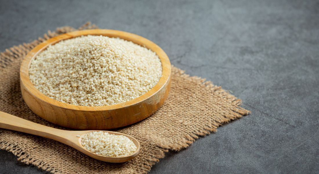 Uma tigela de madeira cheia de psyllium, uma fibra alimentar solúvel, sobre um pedaço de tecido rústico marrom. Ao lado, uma colher de madeira também contendo psyllium. O fundo é uma superfície escura e texturizada.