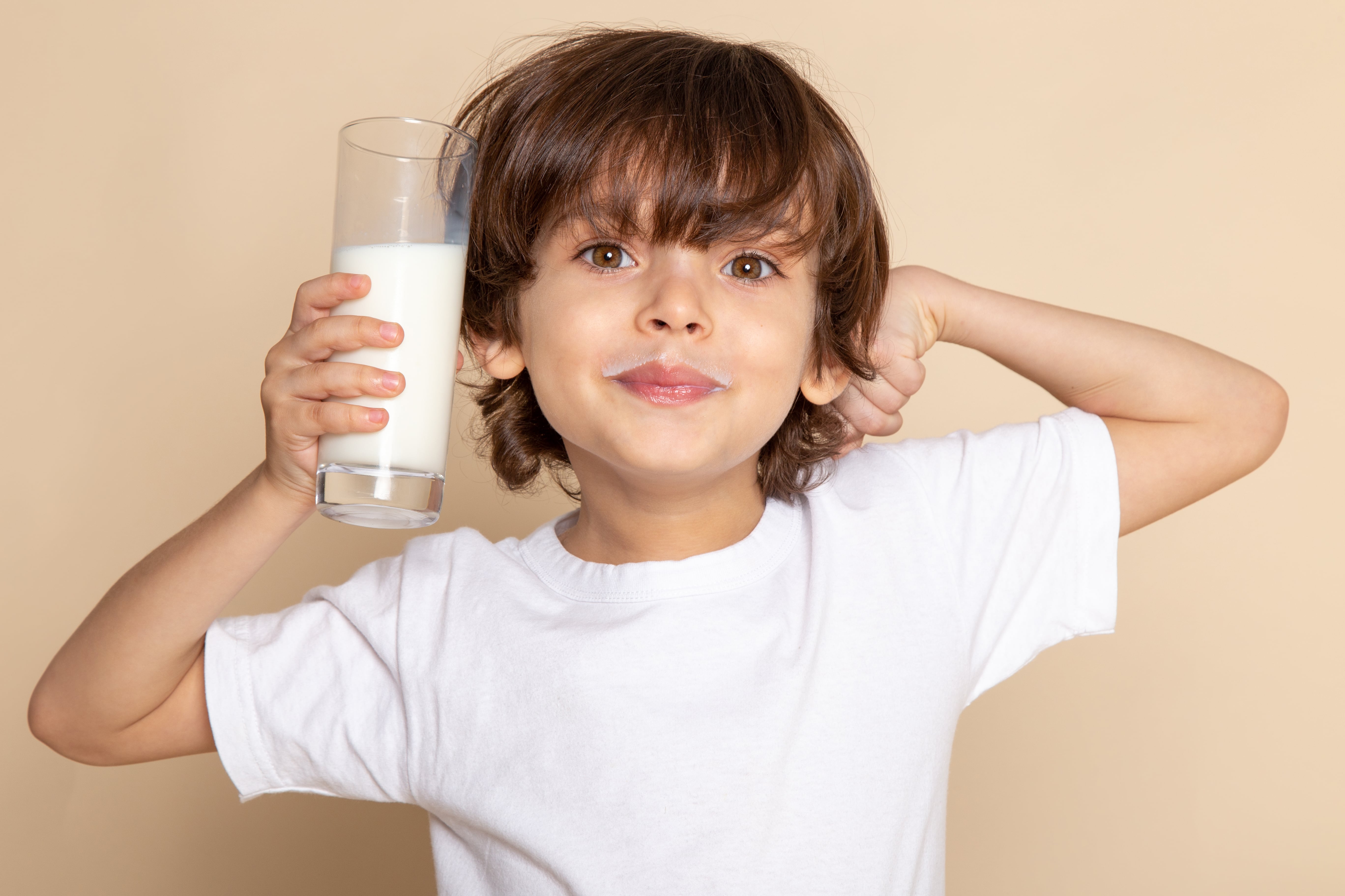 A imagem mostra um menino com cabelo castanho e olhos expressivos, vestindo uma camiseta branca, segurando um copo de leite na mão direita. Ele sorri para a câmera com um bigode de leite nos lábios e posiciona o braço esquerdo dobrado atrás da cabeça. O fundo é bege, e a cena transmite uma sensação de alegria e leveza.