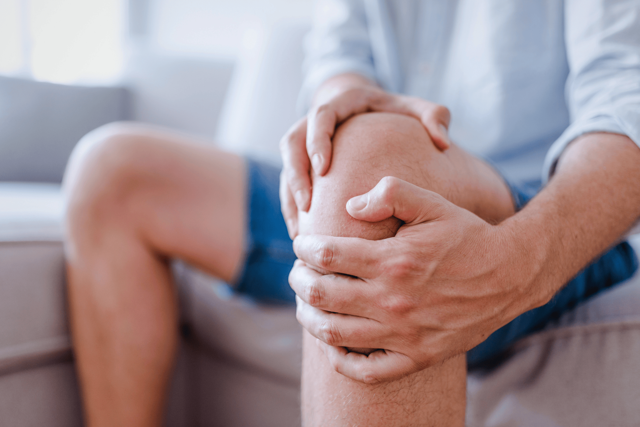 Um close-up de um homem sentado, vestindo uma camisa social clara e bermuda jeans. Ele está com as duas mãos envolvendo o joelho direito, com os dedos pressionando a pele, sugerindo uma tentativa de aliviar uma dor aguda ou crônica.