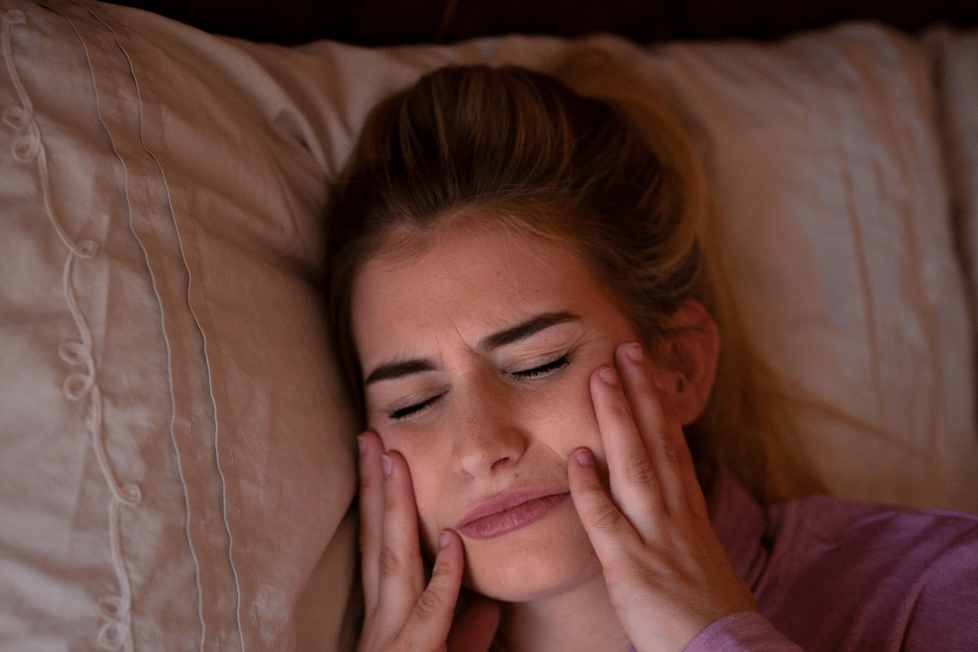 Mulher deitada na cama com expressão de dor, pressionando os dois lados do rosto com as mãos, ilustrando sintomas de DTM (disfunção temporomandibular), como dor na face, tensão na mandíbula e desconforto ao abrir a boca.