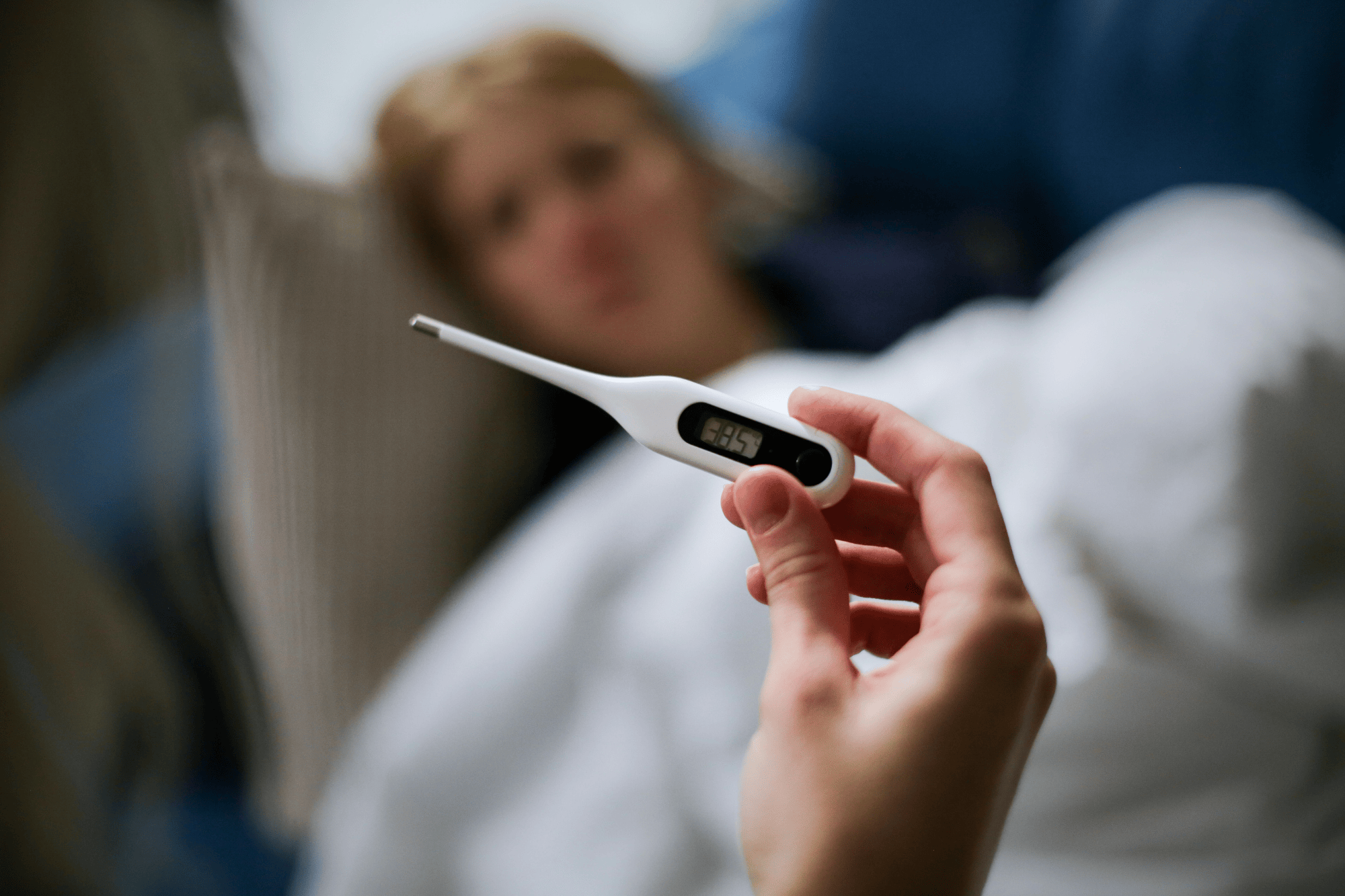 Pessoa deitada na cama, aparentando estar doente, enquanto segura um termômetro digital que marca febre (aproximadamente 38,5 °C), sugerindo quadro de mal-estar ou infecção.