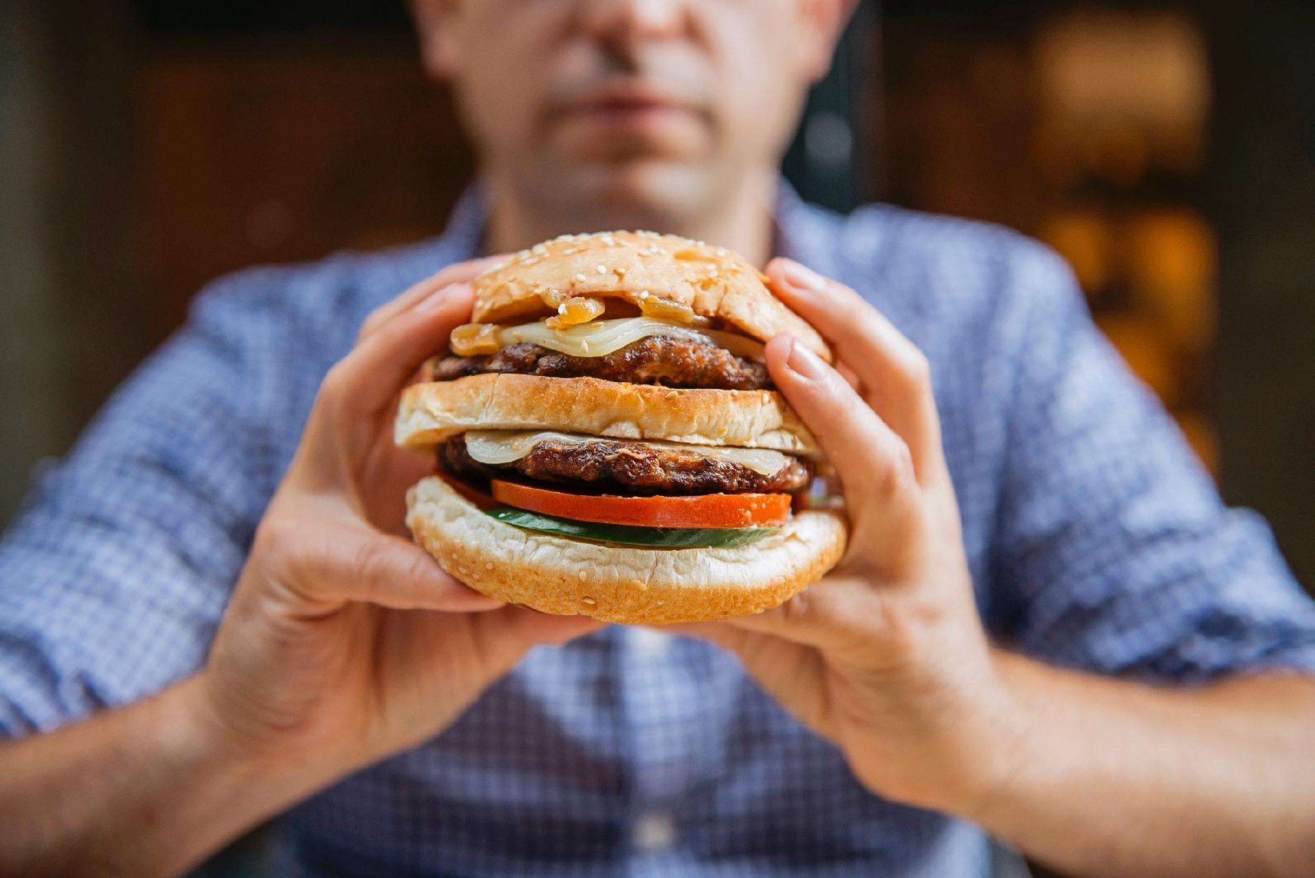 A imagem mostra um homem segurando um hambúrguer grande com ambas as mãos.