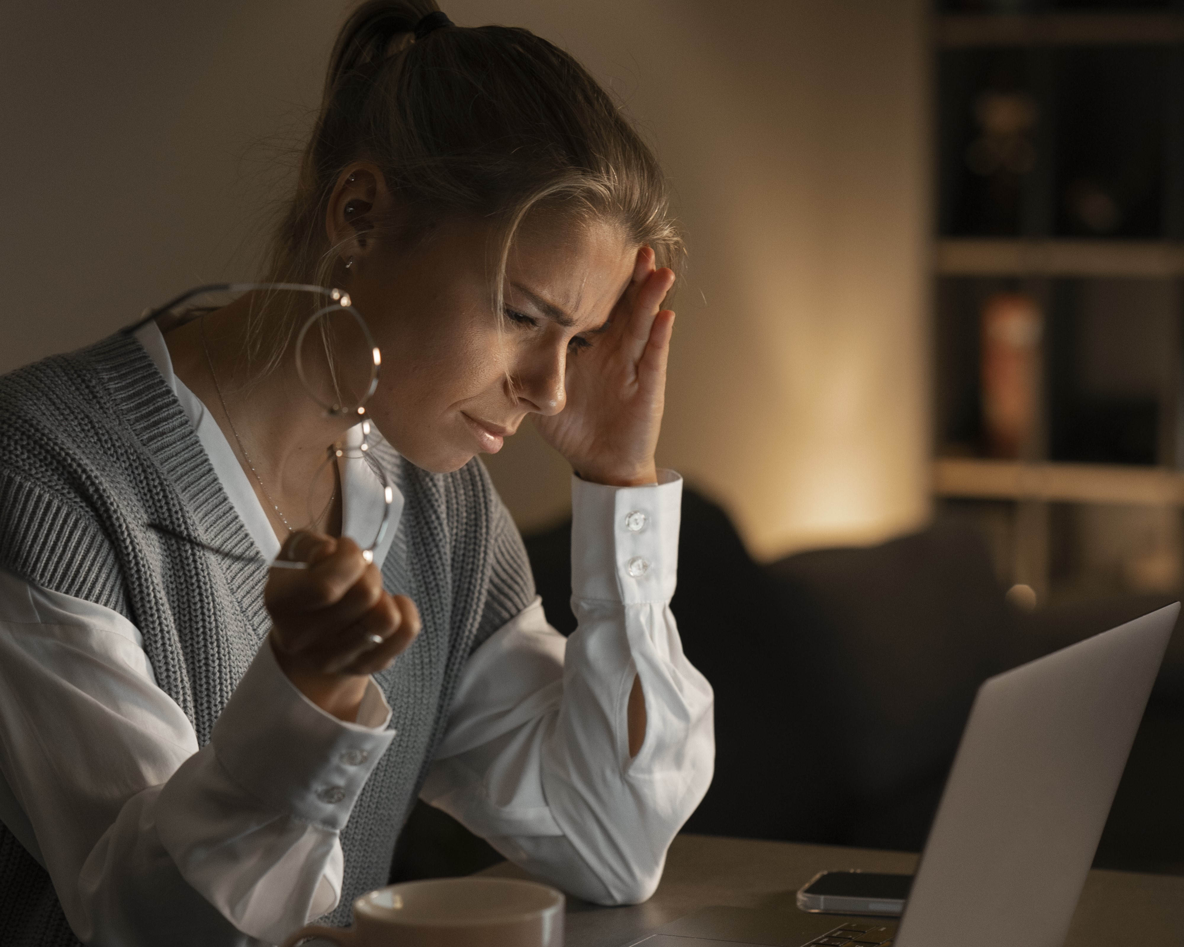 Mulher jovem aparentando cansaço extremo, sentada à frente de um laptop à noite, com expressão de fadiga, segurando os óculos em uma mão e apoiando a cabeça com a outra, sugerindo que dormiu pouco e está sobrecarregada de trabalho. Há uma xícara de café e cadernos sobre a mesa, reforçando o ambiente de esforço contínuo.