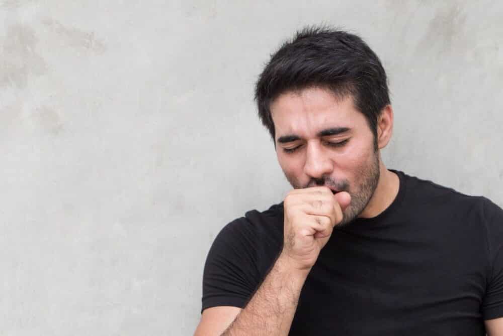 A imagem mostra um homem de barba, vestindo uma camiseta preta, tossindo com a mão fechada sobre a boca. Ele aparenta estar com desconforto respiratório, possivelmente devido a uma infecção pulmonar, como a pneumonia viral. O fundo da imagem é uma parede cinza.